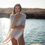 woman wearing lightweight mersea nellie wrap in neutral moonstone color draped around shoulders and arms standing in front of water