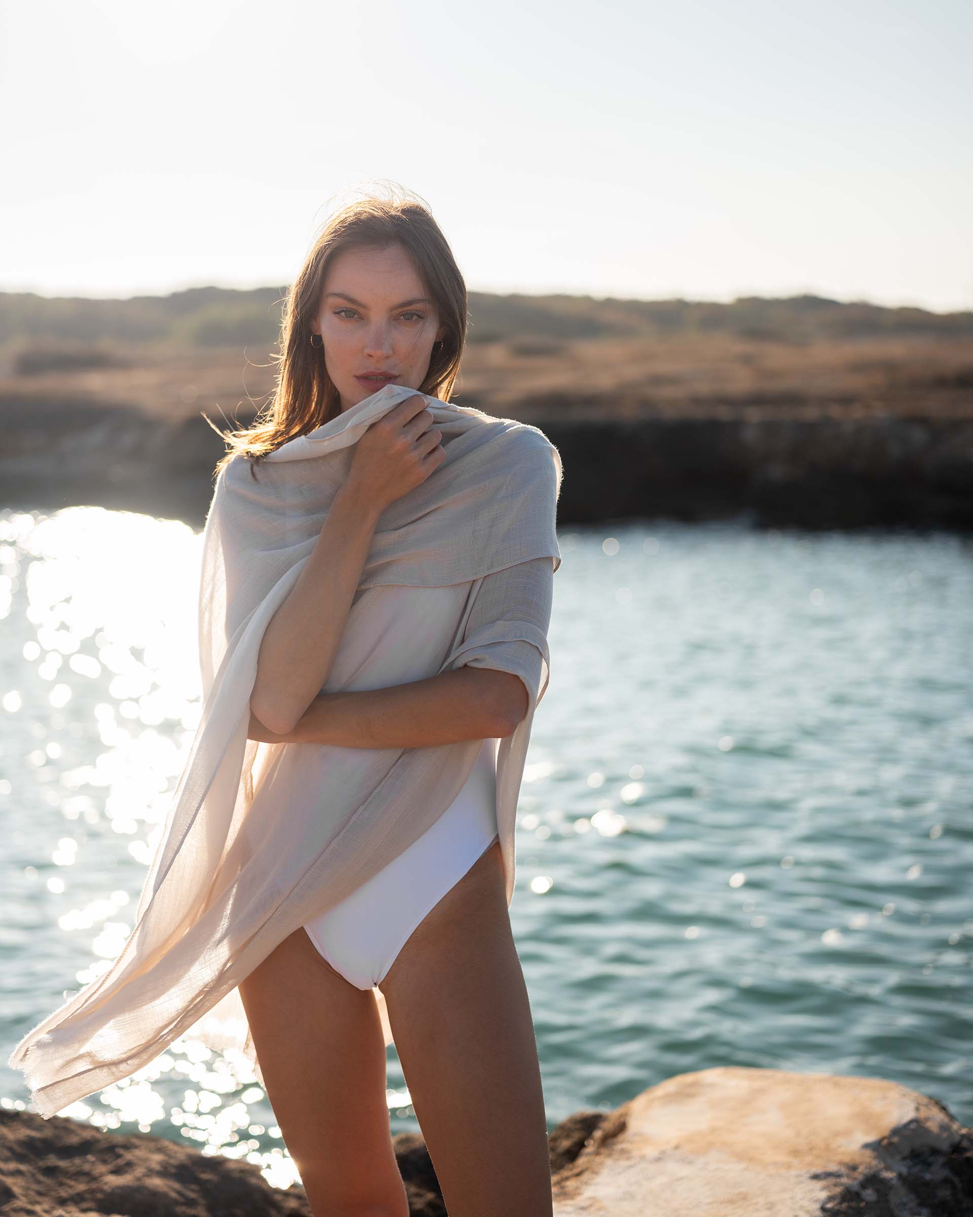 woman wearing lightweight mersea nellie wrap in neutral moonstone color draped around shoulders and arms standing in front of water