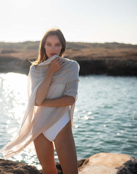 woman wearing lightweight mersea nellie wrap in neutral moonstone color draped around shoulders and arms standing in front of water