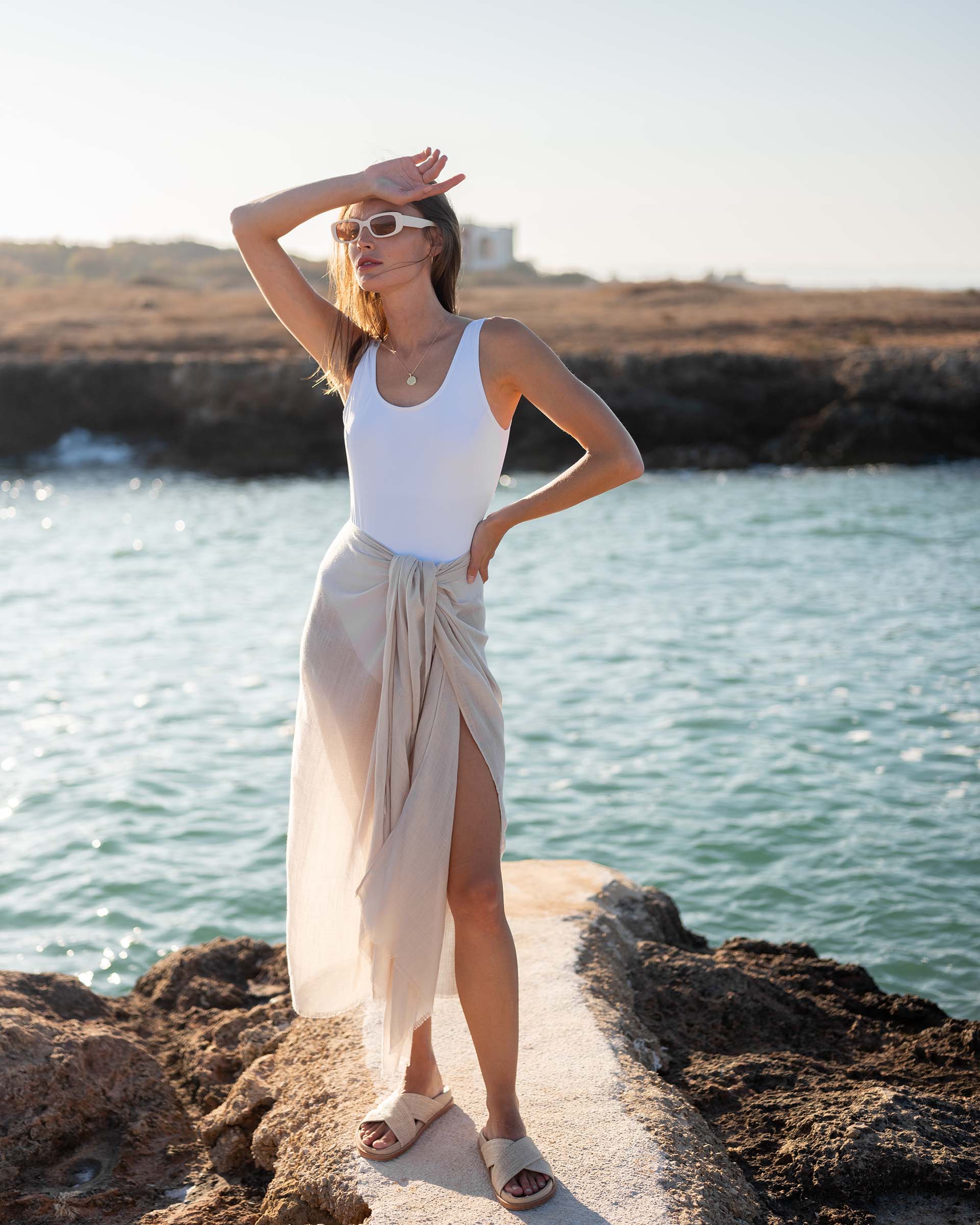 woman wearing lightweight mersea nellie wrap in neutral moonstone color wrapped around waist standing in front of body of water