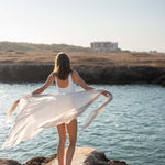 woman holding lightweight mersea nellie wrap in neutral moonstone color flowing in the wind near body of water