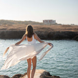 woman holding lightweight mersea nellie wrap in neutral moonstone color flowing in the wind near body of water