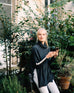 woman wearing mersea newport sweater in storm grey with light salt accent stripe down lower sleeve with a split front turtleneck and ribbed sleeves holding phone outside 