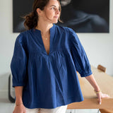 Woman wearing a blue blouse standing indoors with a painting in the background