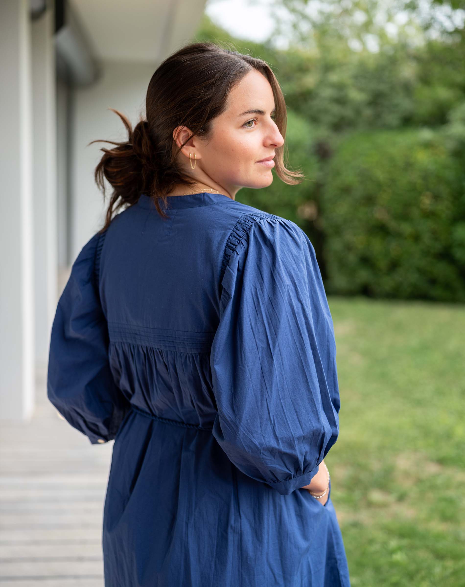 Woman wearing a blue dress standing outdoors with greenery in the background
