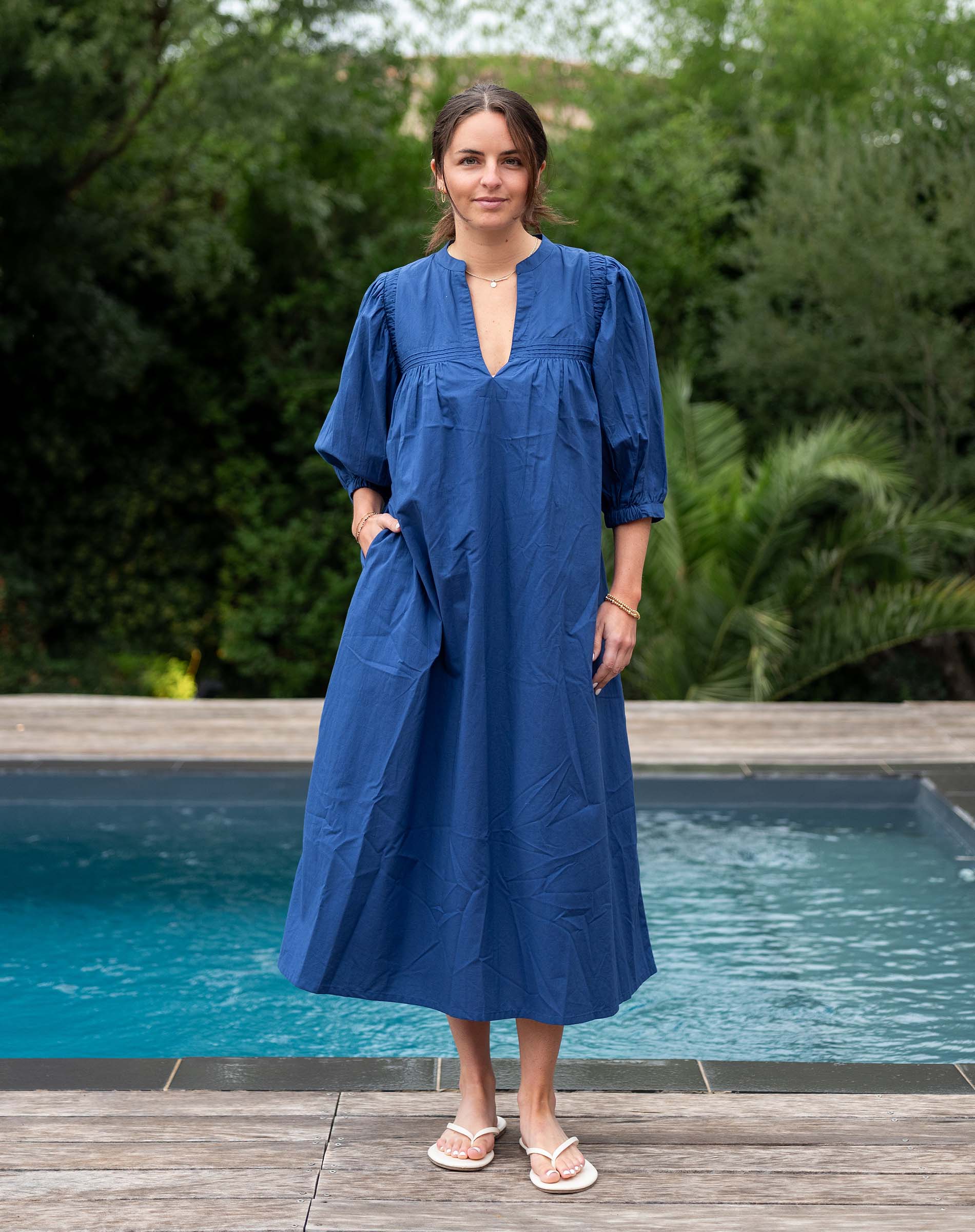 Woman in a blue dress standing by a pool with greenery in the background