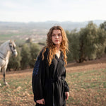 female wearing Mersea black embroidered maxi dress with tassel ties in a field in front of a horse