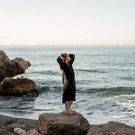 female wearing Mersea black embroidered maxi dress with tassel ties standing on a rock in front of water