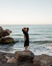female wearing Mersea black embroidered maxi dress with tassel ties standing on a rock in front of water