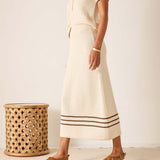 Person wearing a cream-colored outfit with a striped skirt standing next to a wooden stool.