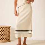 Person wearing a long, light-colored skirt with dark trim against a plain background