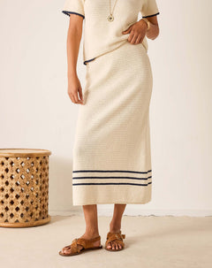 Person wearing a long, light-colored skirt with dark trim against a plain background