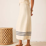 Person wearing a long, light-colored dress with a striped pattern, standing next to a woven stool.