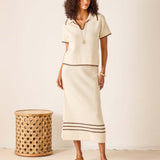 Woman wearing a cream-colored outfit with a skirt, standing next to a woven stool against a white background.