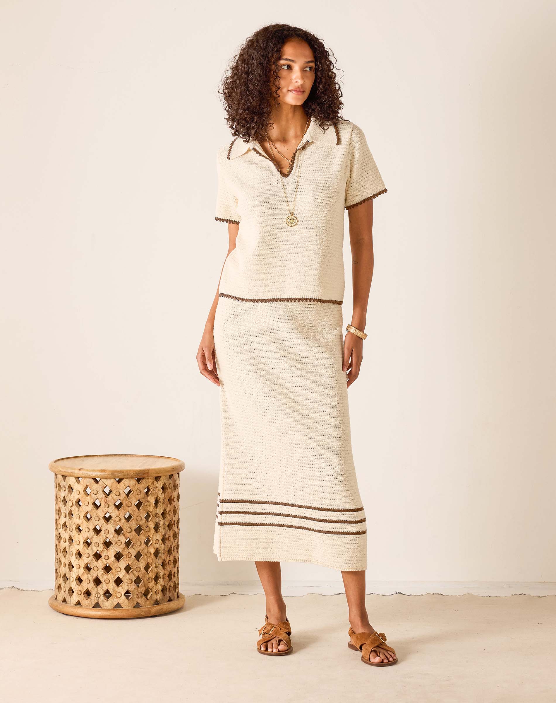 Woman wearing a cream-colored outfit with a skirt, standing next to a woven stool against a white background.