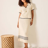 Woman wearing a cream-colored outfit with black trim standing next to a woven stool against a white background