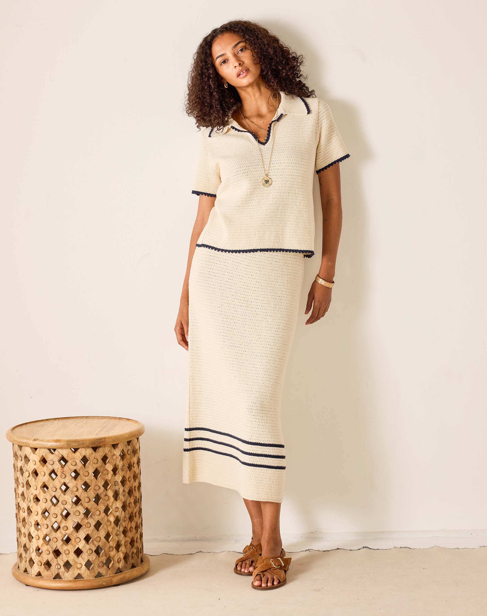 Woman wearing a cream-colored outfit with black trim standing next to a woven stool against a white background