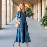 Woman in a blue dress standing in an architectural hallway with columns and plants.