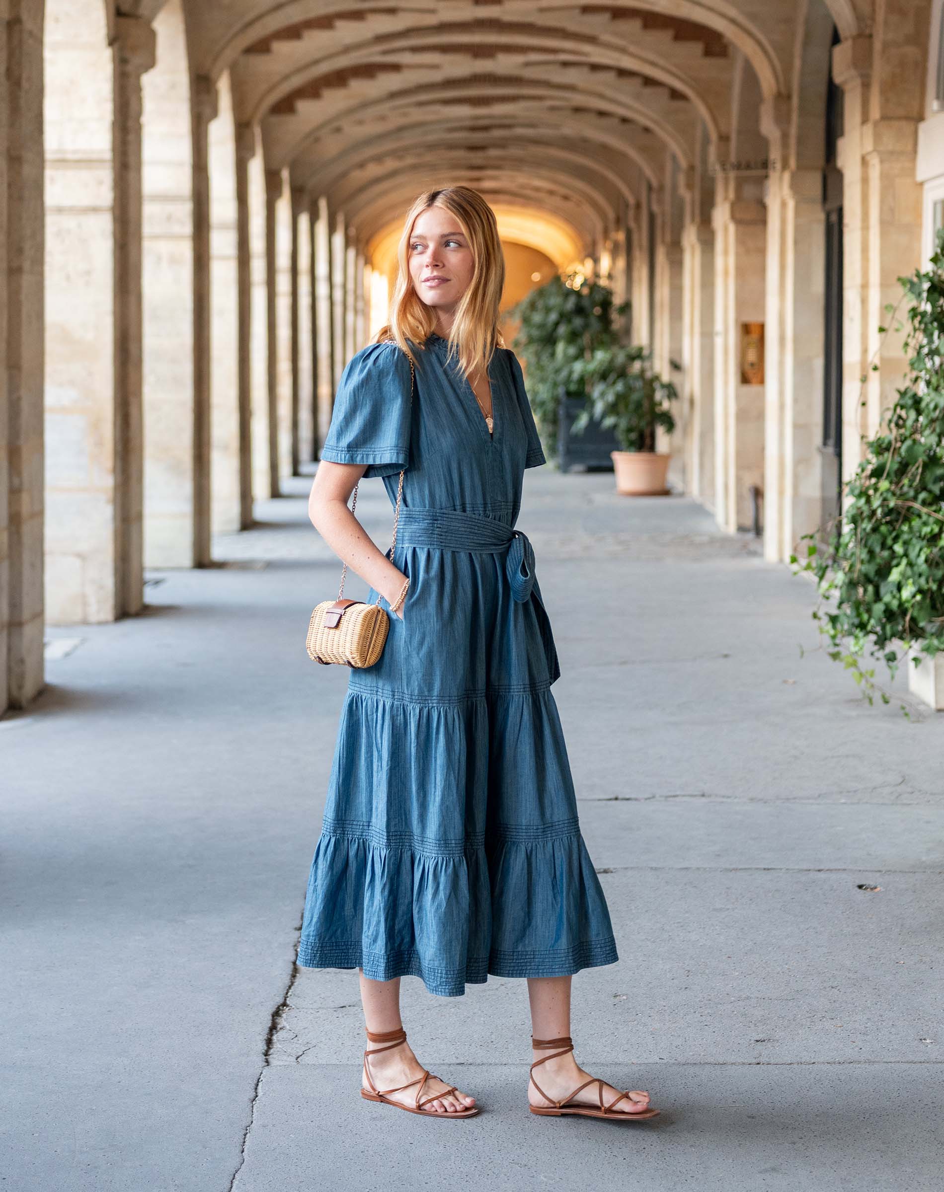 Woman in a blue dress standing in an architectural hallway with columns and plants.