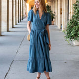 Woman in a blue dress standing in an outdoor setting with columns and plants.