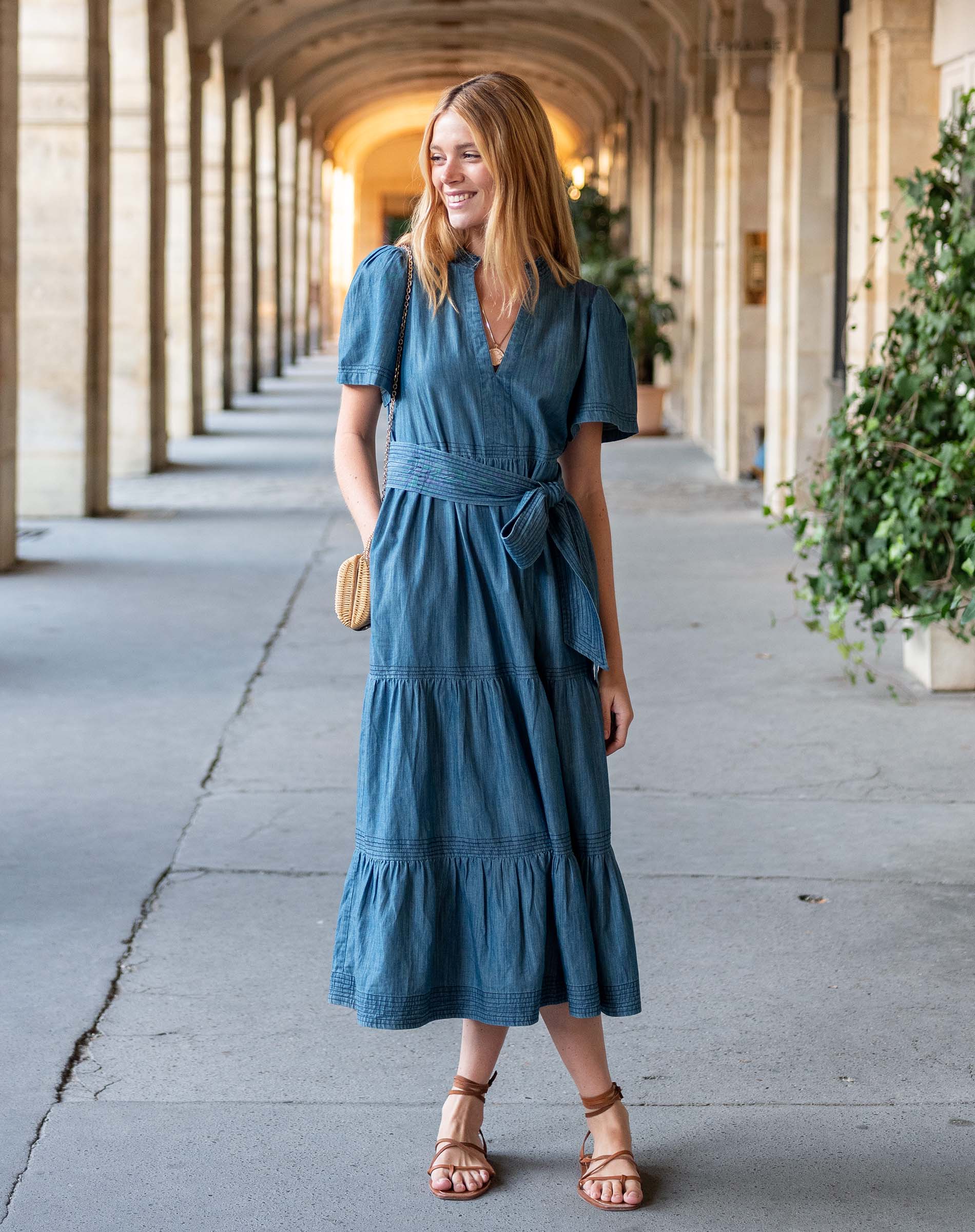 Woman in a blue dress standing in an outdoor setting with columns and plants.