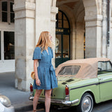 Woman in a blue dress standing next to a vintage car under arches of a building.