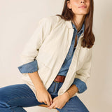 Woman wearing a beige jacket over a denim shirt and jeans, sitting on a chair against a plain background.