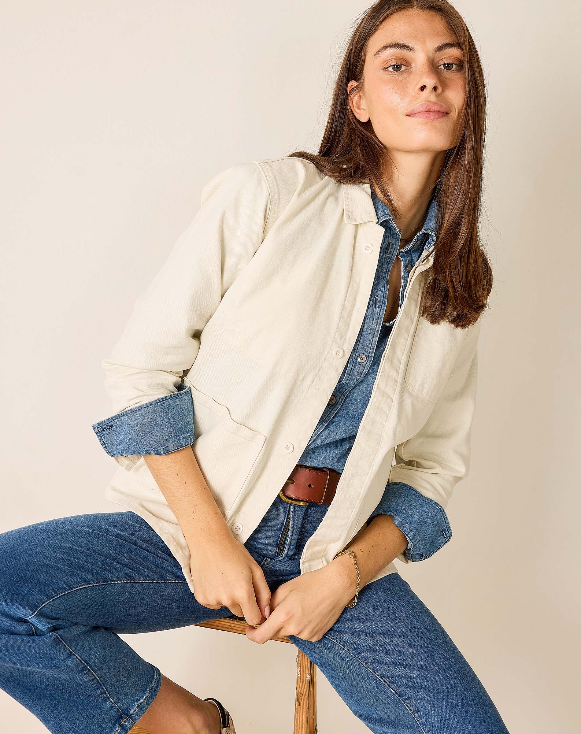 Woman wearing a beige jacket over a denim shirt and jeans, sitting on a chair against a plain background.