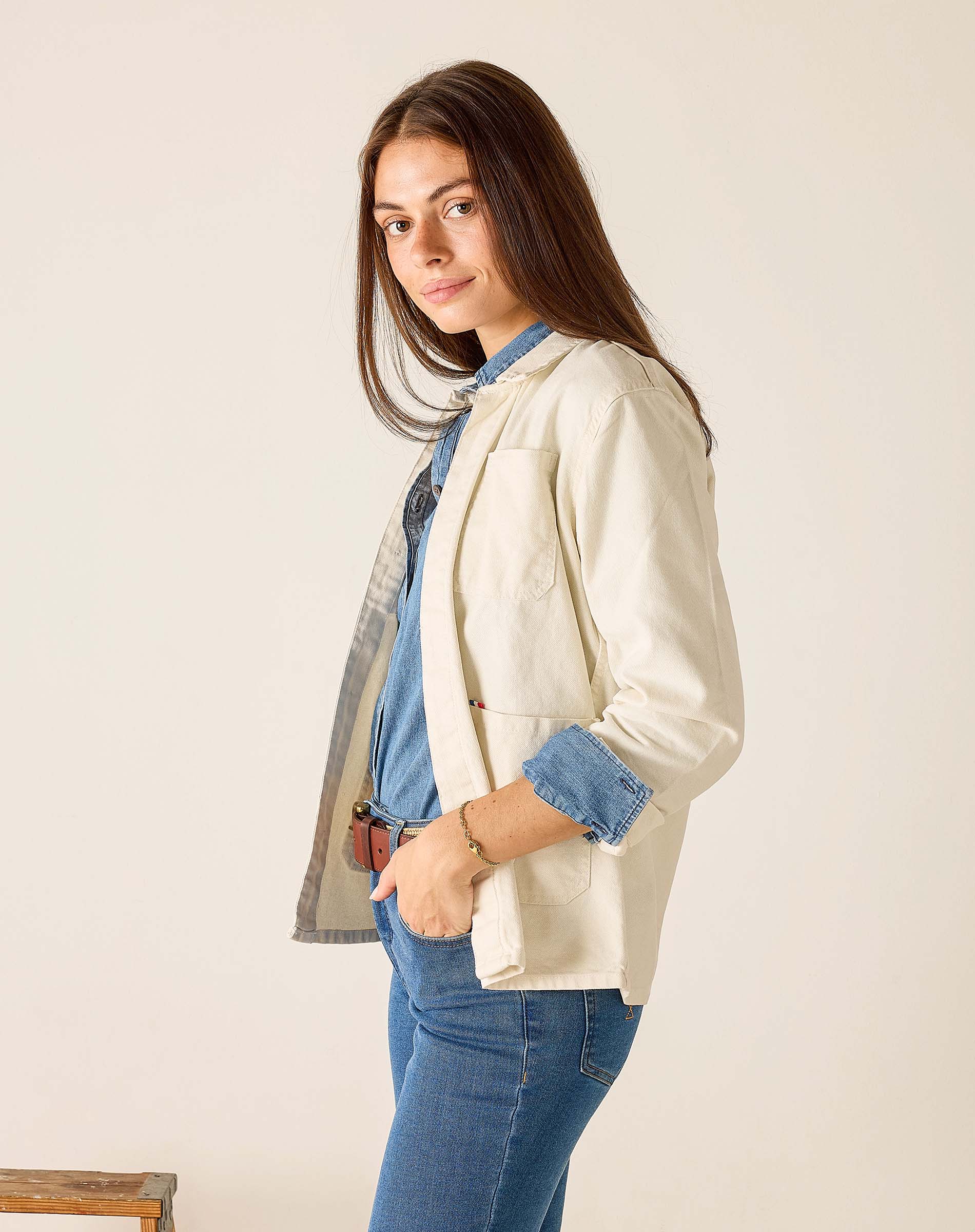 Woman wearing a cream blazer over a denim shirt and jeans on a plain background