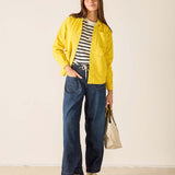 Woman wearing a yellow jacket, striped shirt, and blue jeans against a plain background