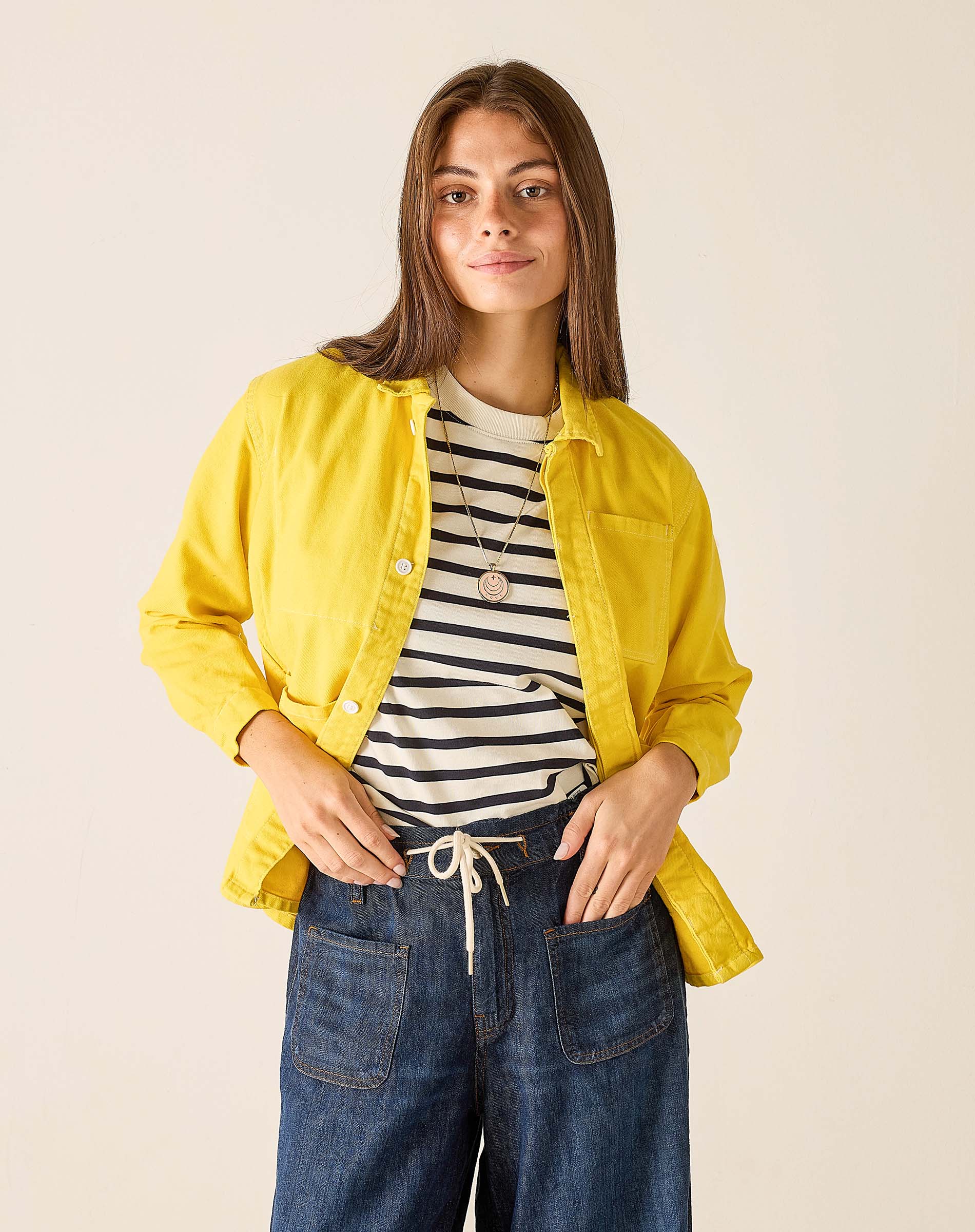 Woman wearing a yellow jacket, striped shirt, and blue jeans on a beige background