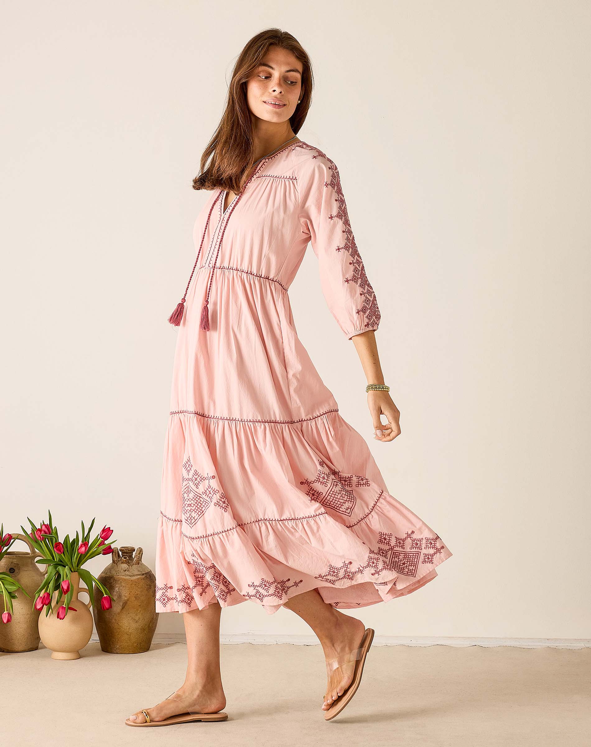 Woman wearing a pink dress with floral embroidery standing in front of a neutral background.