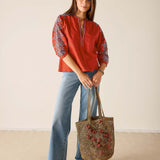 Woman wearing a red top and blue jeans, holding a woven bag with floral designs, on a beige background.