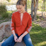 Woman wearing a red embroidered blouse and blue jeans sitting on a rock outdoors.