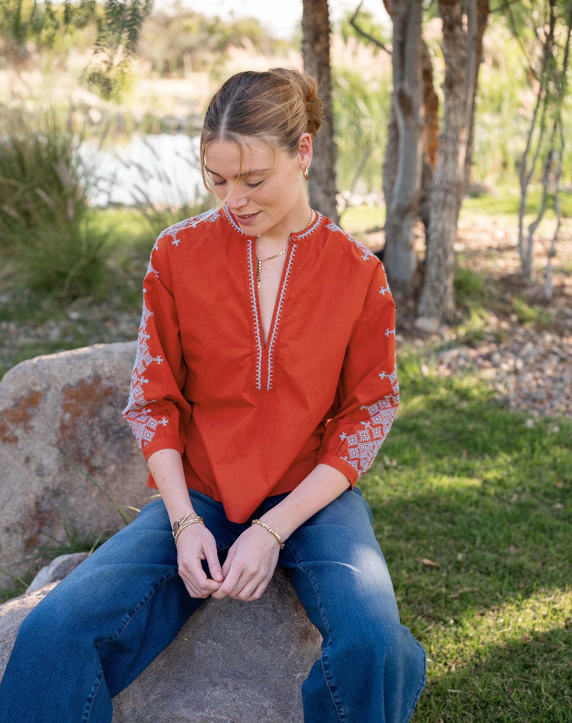Woman wearing a red embroidered blouse and blue jeans sitting on a rock outdoors.