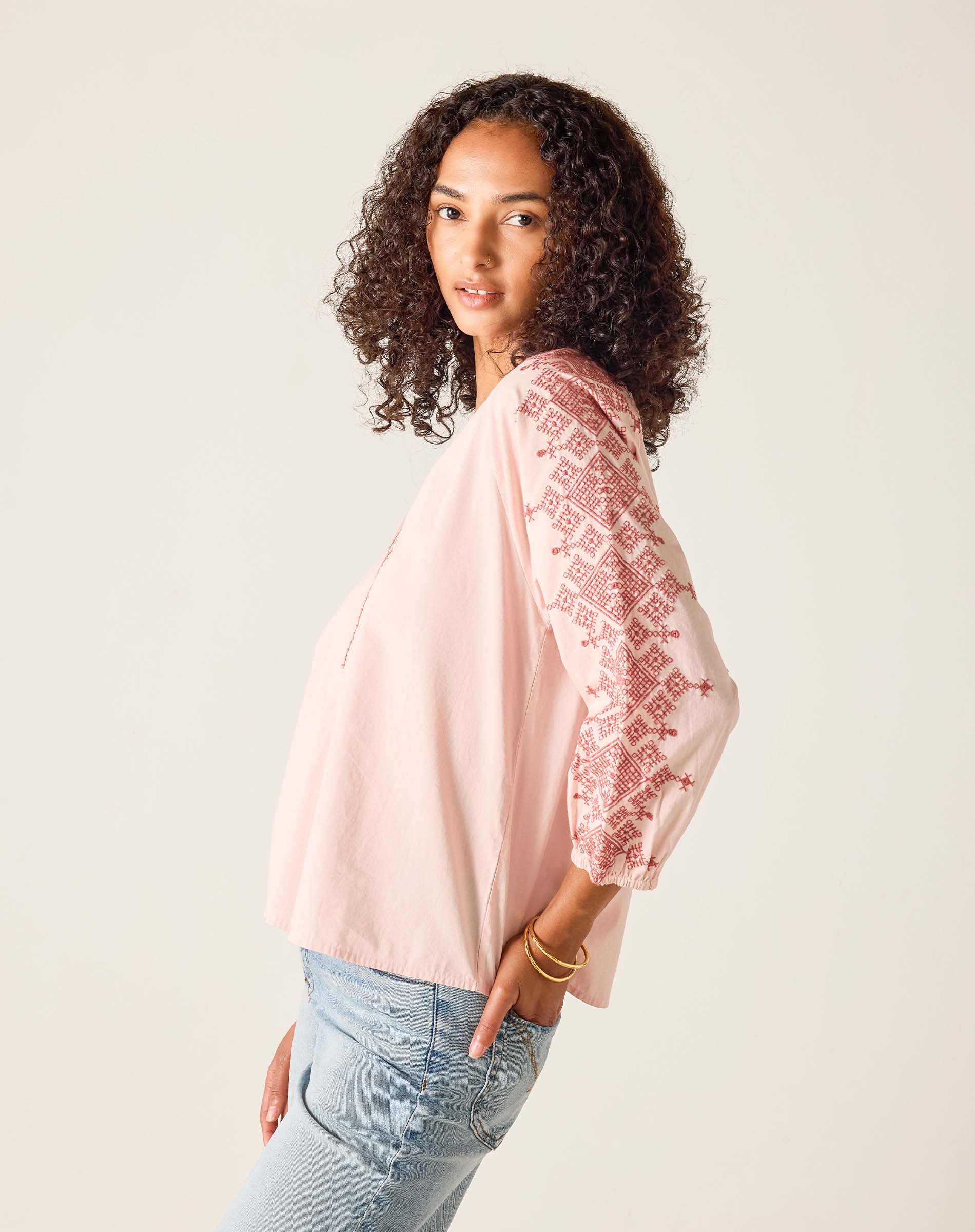 Woman wearing a light pink blouse with embroidery and light blue jeans against a beige background