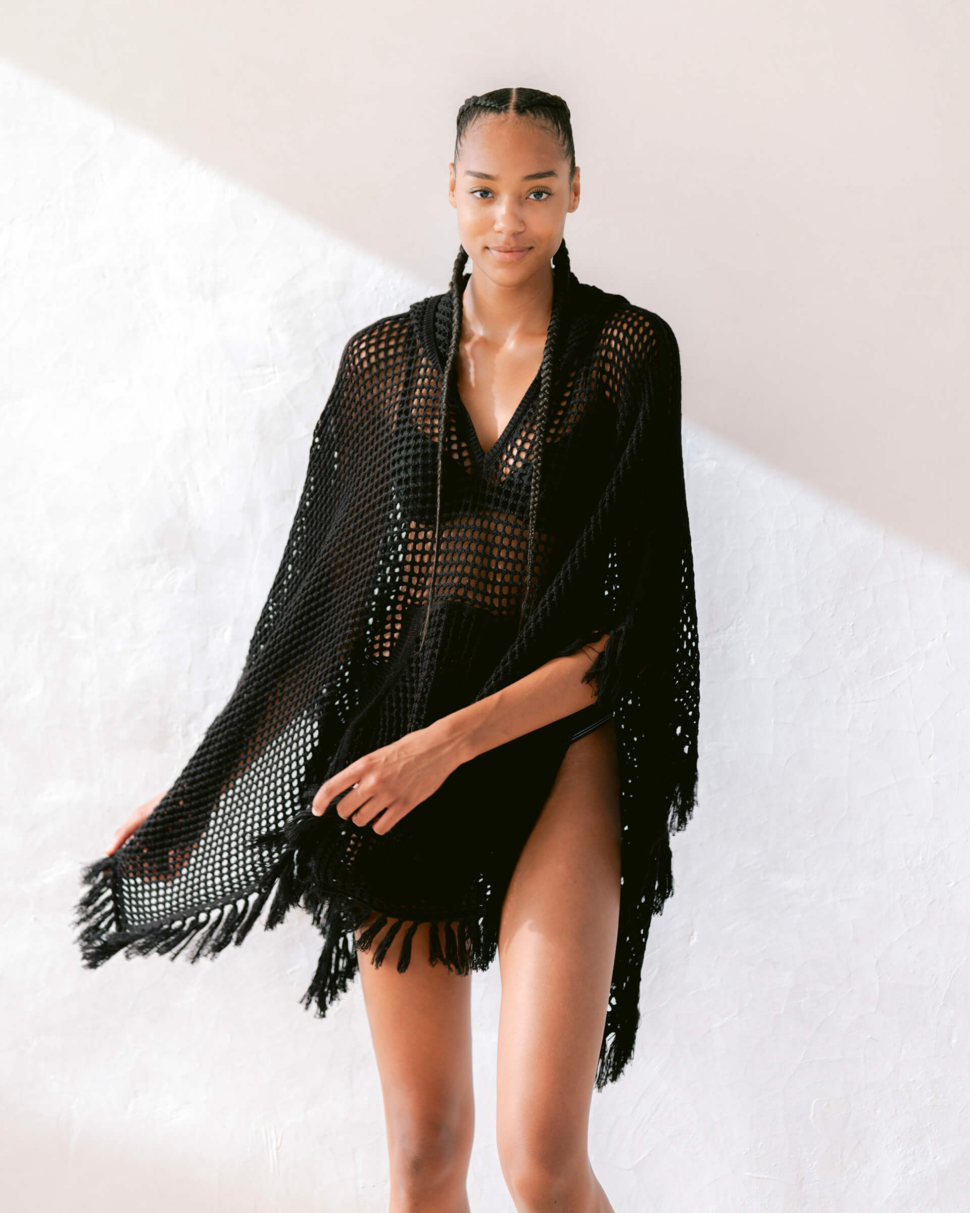 female wearing black fringe crochet-knit hoodie swaying on a white wall