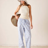 Woman wearing a white sleeveless top and light blue striped pants, holding a straw bag against a plain background.