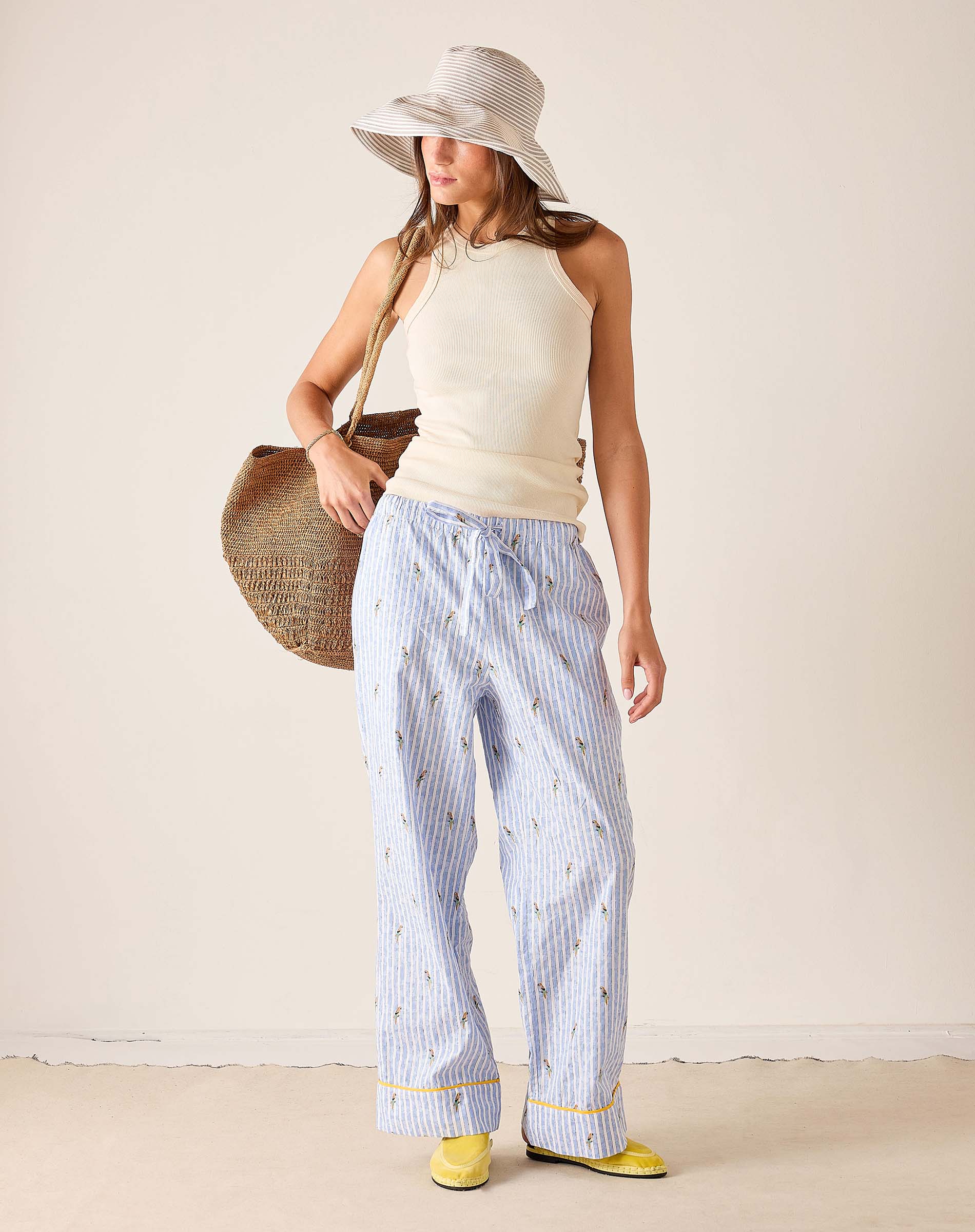 Woman wearing a white sleeveless top and light blue striped pants, holding a straw bag against a plain background.
