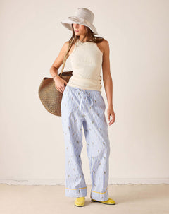 Woman wearing a white sleeveless top and light blue striped pants, holding a straw bag against a plain background.