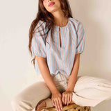 Woman wearing a striped blouse sitting on a woven stool against a plain background