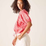 Woman wearing a red patterned top and white pants against a plain background