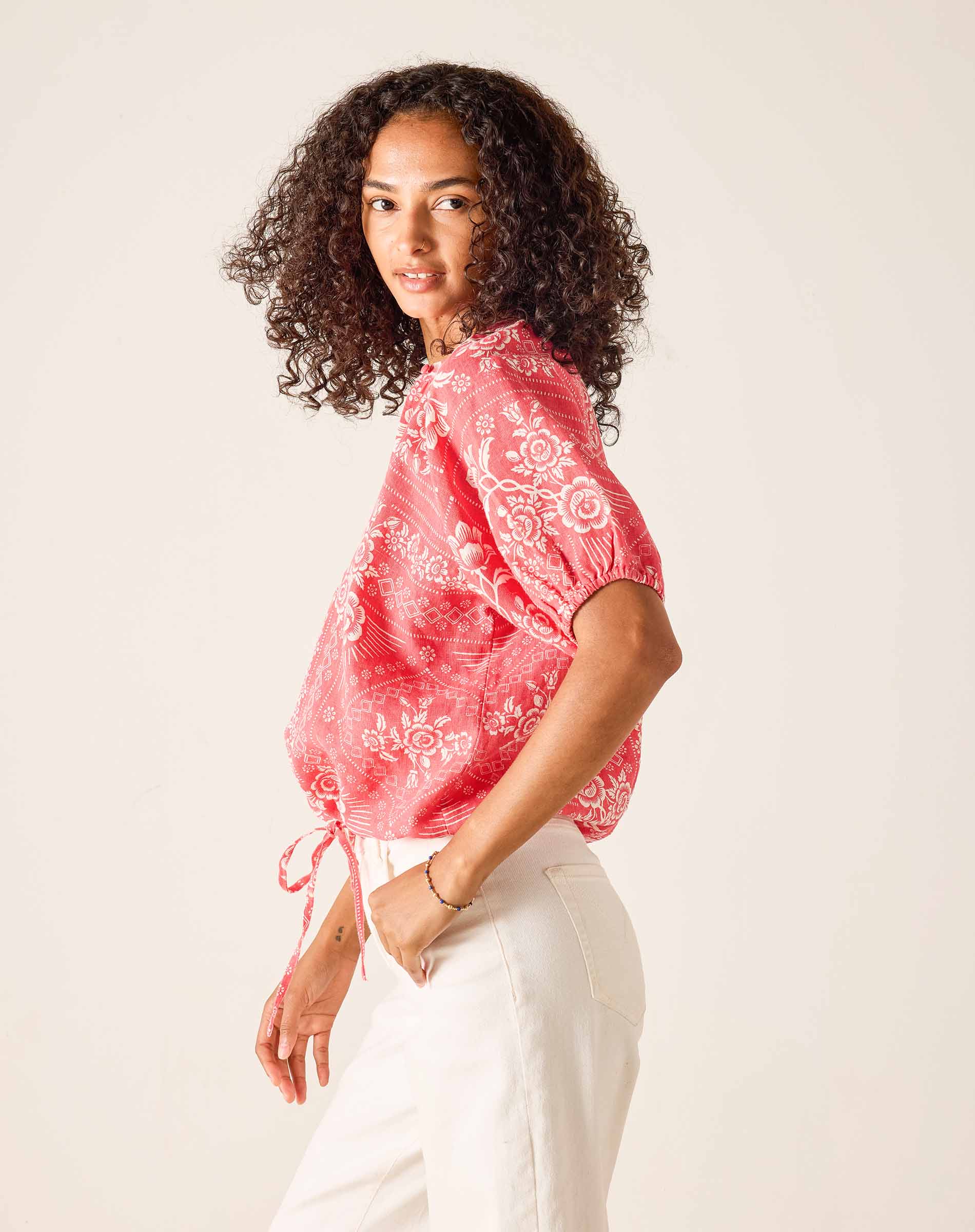 Woman wearing a red patterned top and white pants against a plain background