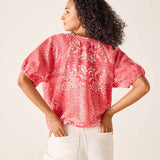 Woman wearing a red patterned blouse with white pants on a plain background