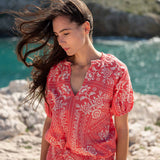 Woman in a red floral dress standing on a rocky shore with water and rocks in the background