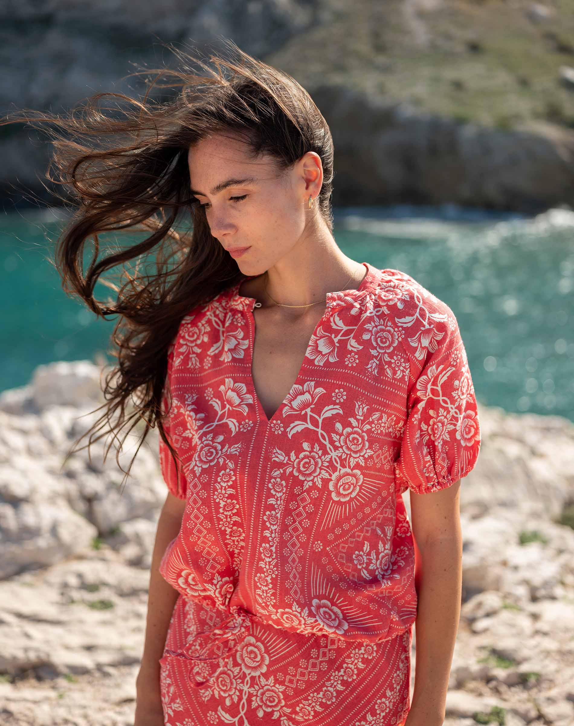 Woman in a red floral dress standing on a rocky shore with water and rocks in the background