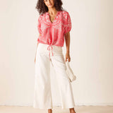 Woman wearing a pink blouse with white patterns and white pants against a plain background