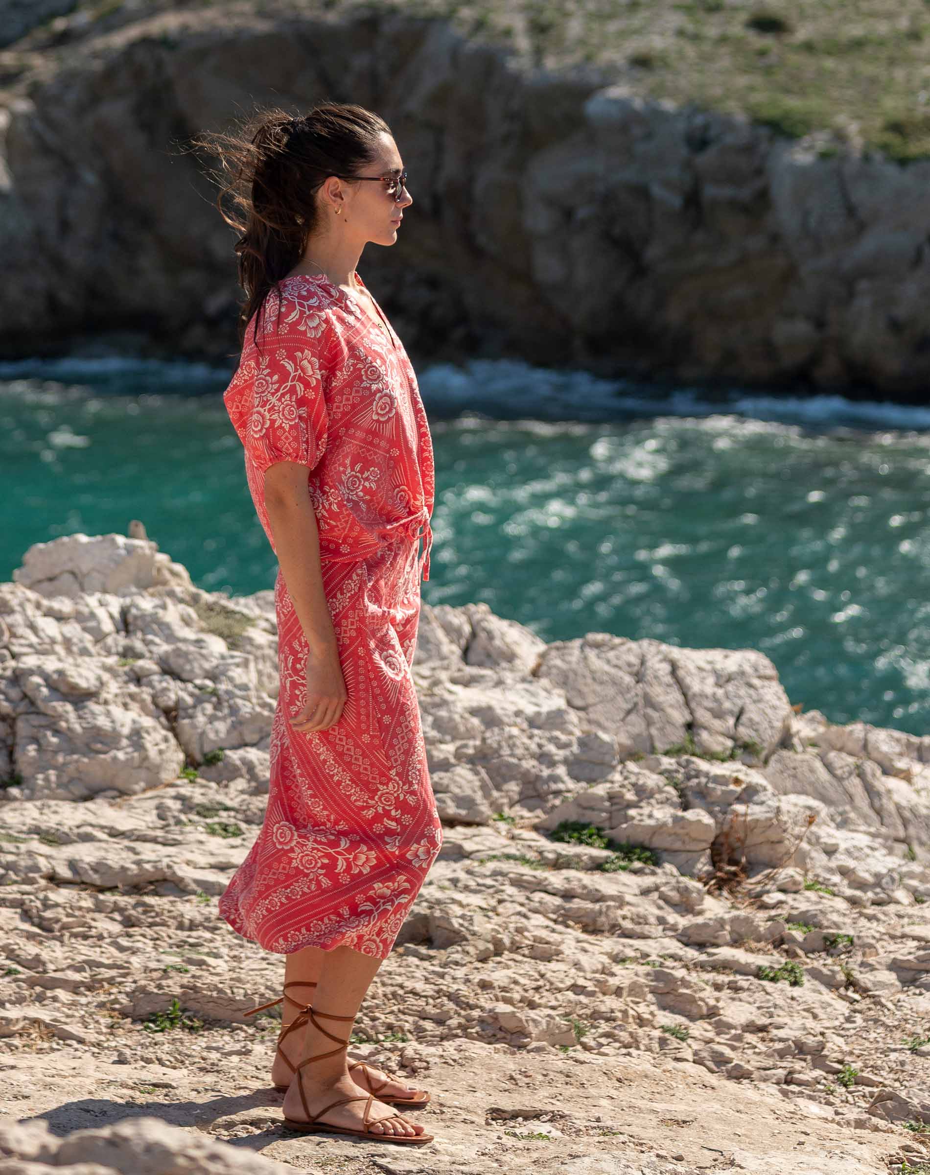 Woman in a red dress standing on rocky cliffs overlooking the ocean.