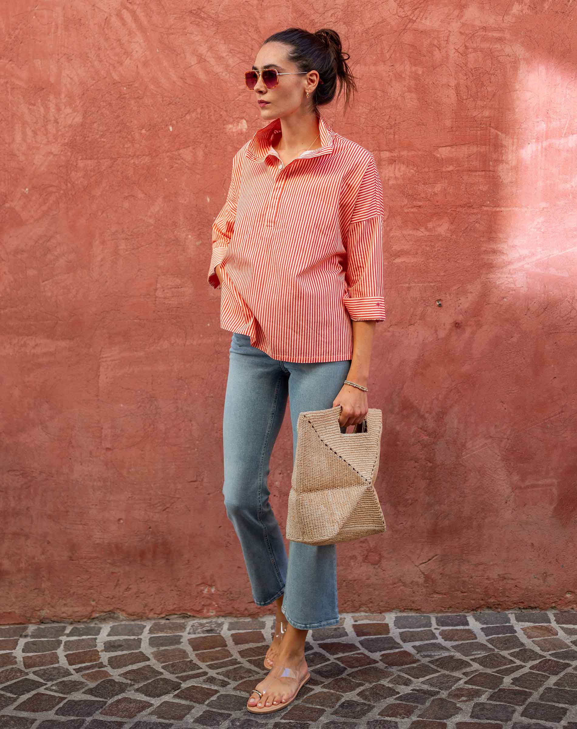 Woman in a pink checkered shirt and blue jeans standing against a textured red wall.