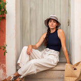 Woman sitting on a step wearing a hat, navy top, and white pants with a straw bag.